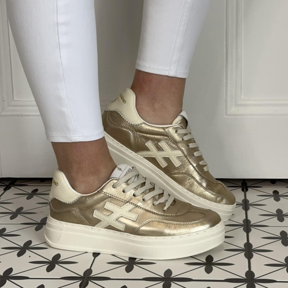 Another Trend Gold sneakers worn with white pants on a patterned floor.