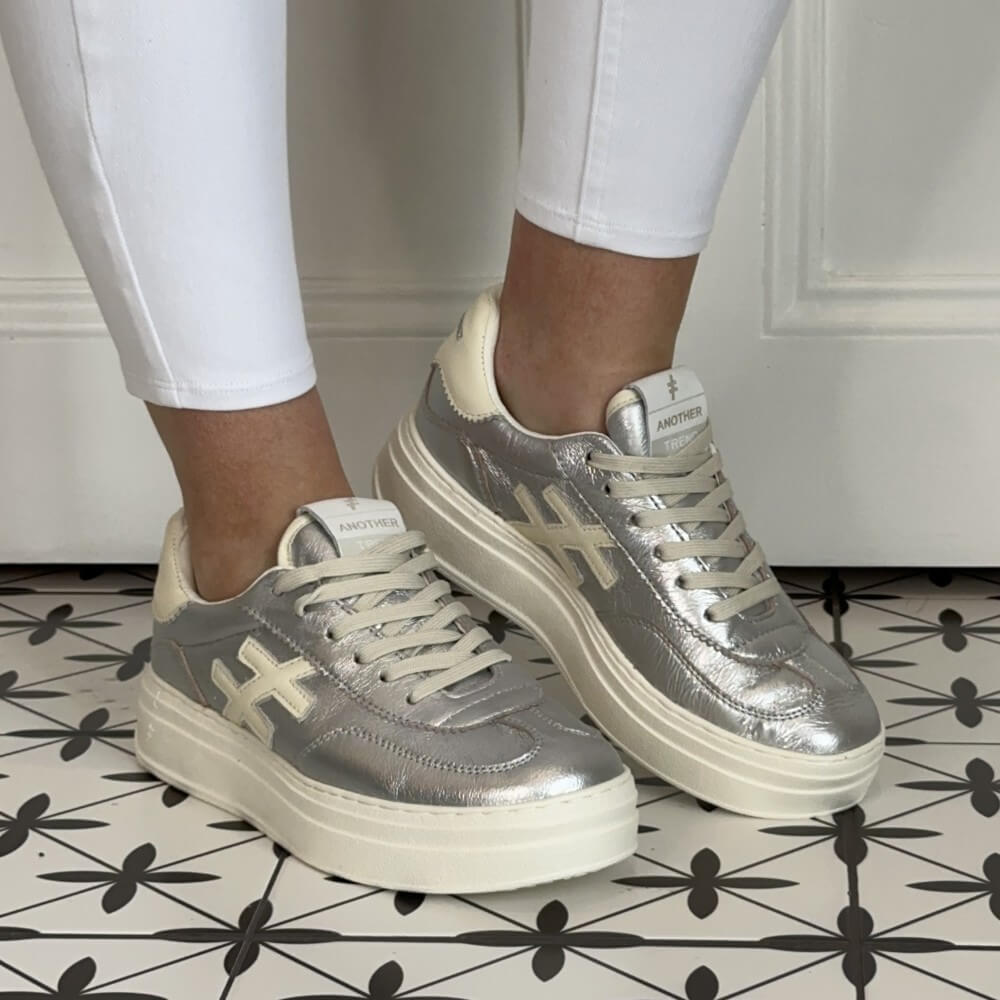 Another Trend Silver and beige sneakers worn with white pants on a patterned floor. 