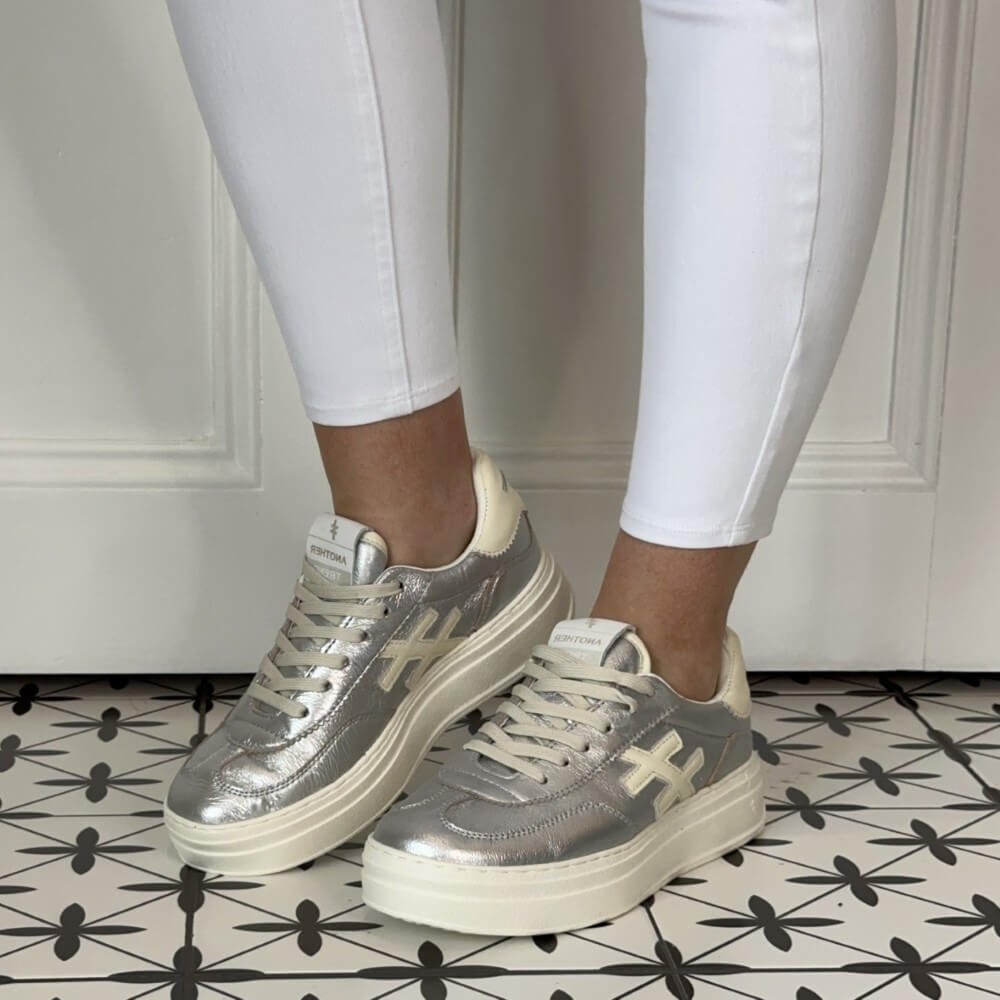 Another Trend Silver and beige sneakers worn with white pants on a patterned floor. 