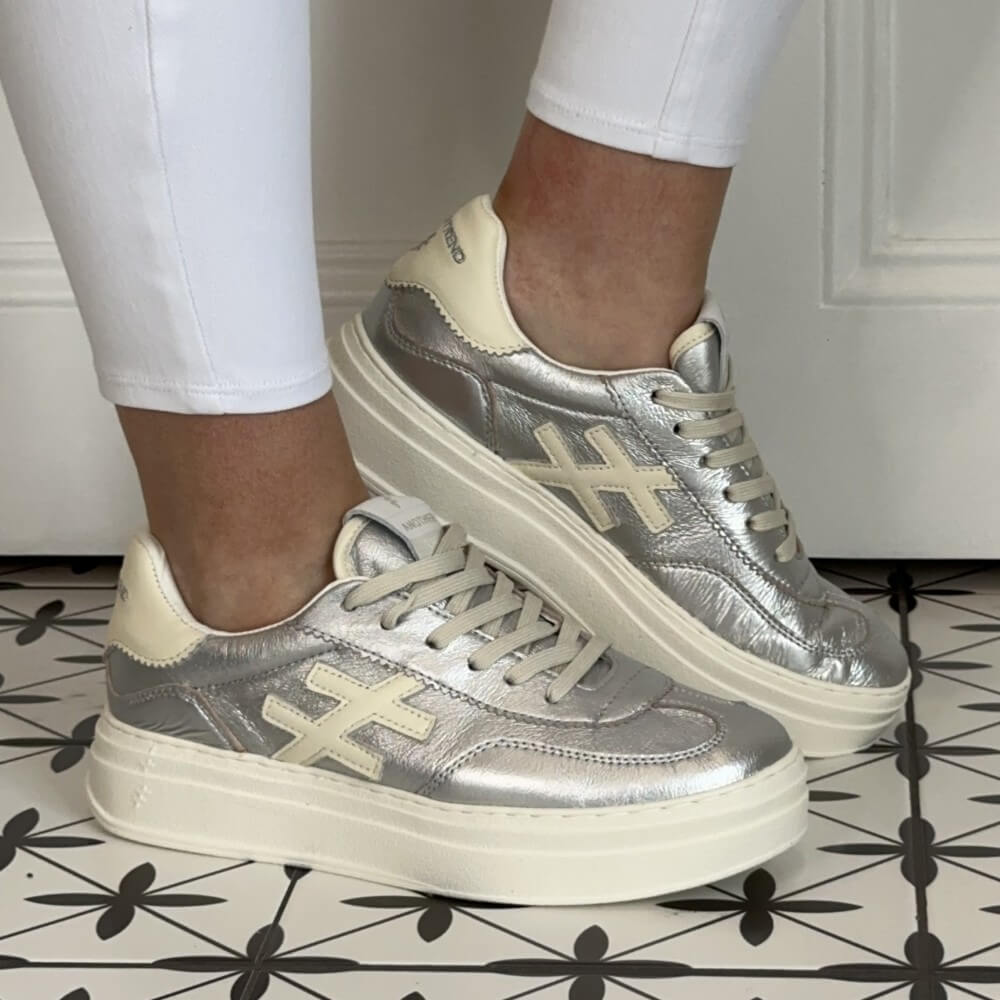 Another Trend Silver and beige sneakers worn with white pants on a patterned floor. 