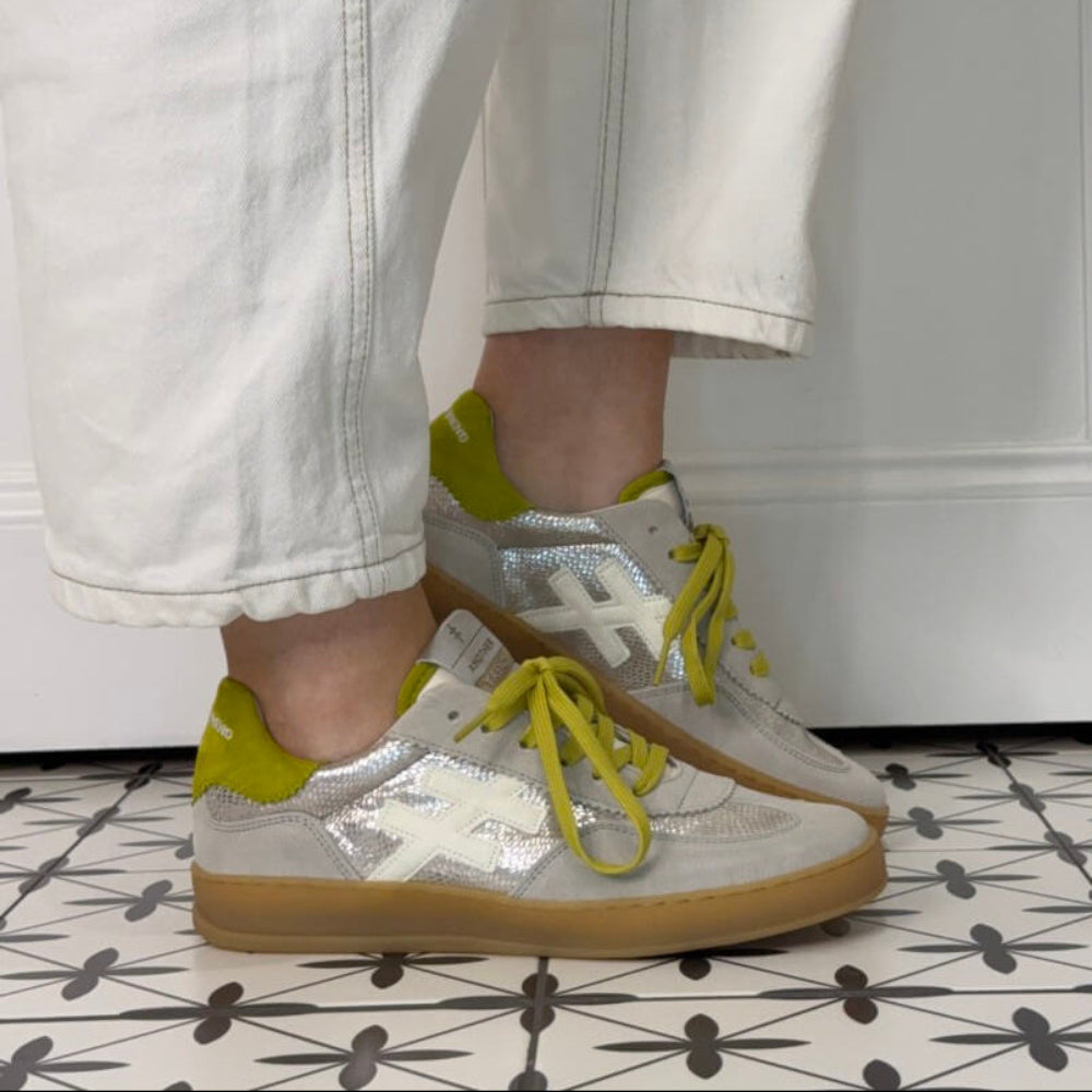 Champagne  sneakers with yellow laces worn by a person on a patterned floor.
