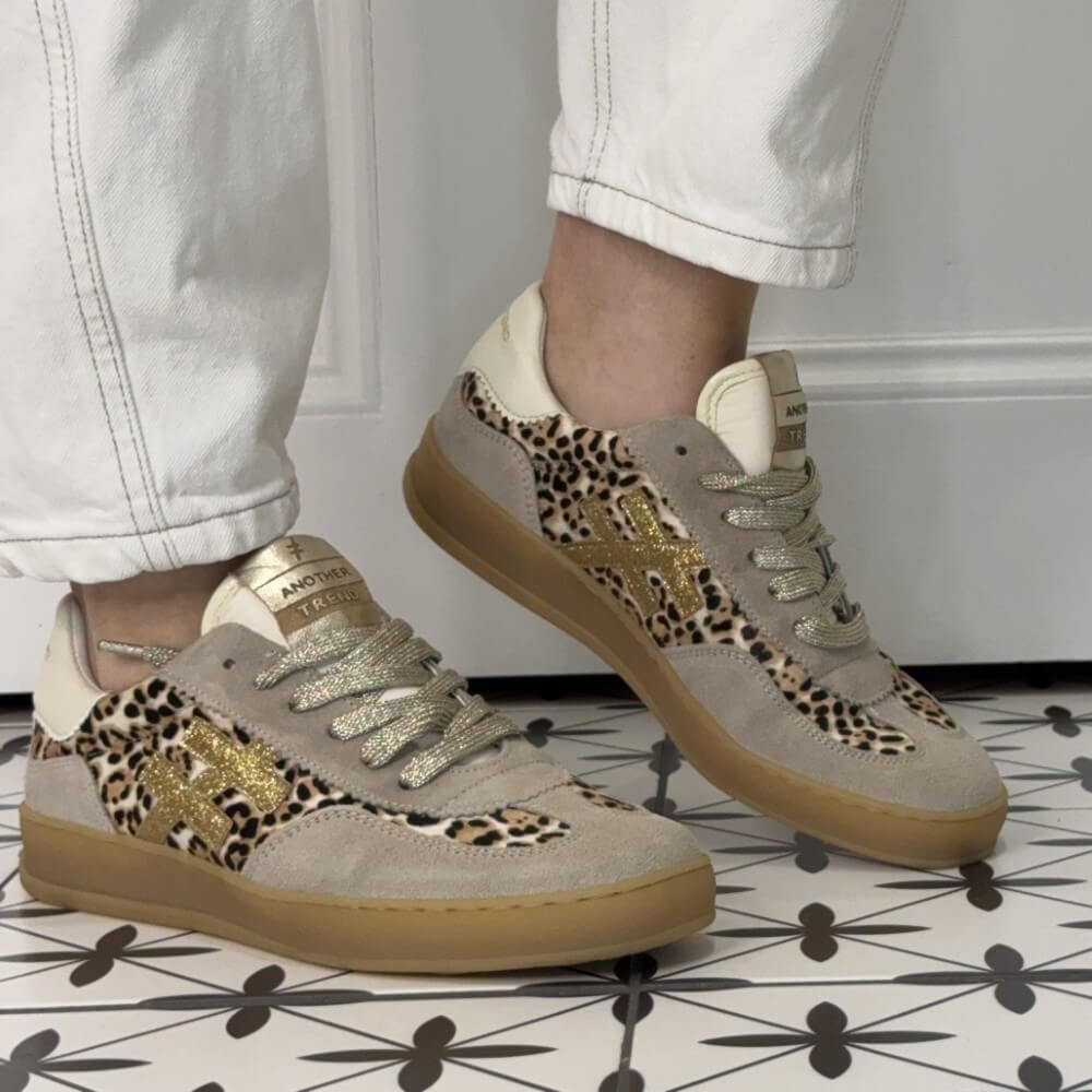 Another Trend Beige sneakers with leopard print and gold accents worn with white pants on a patterned floor.