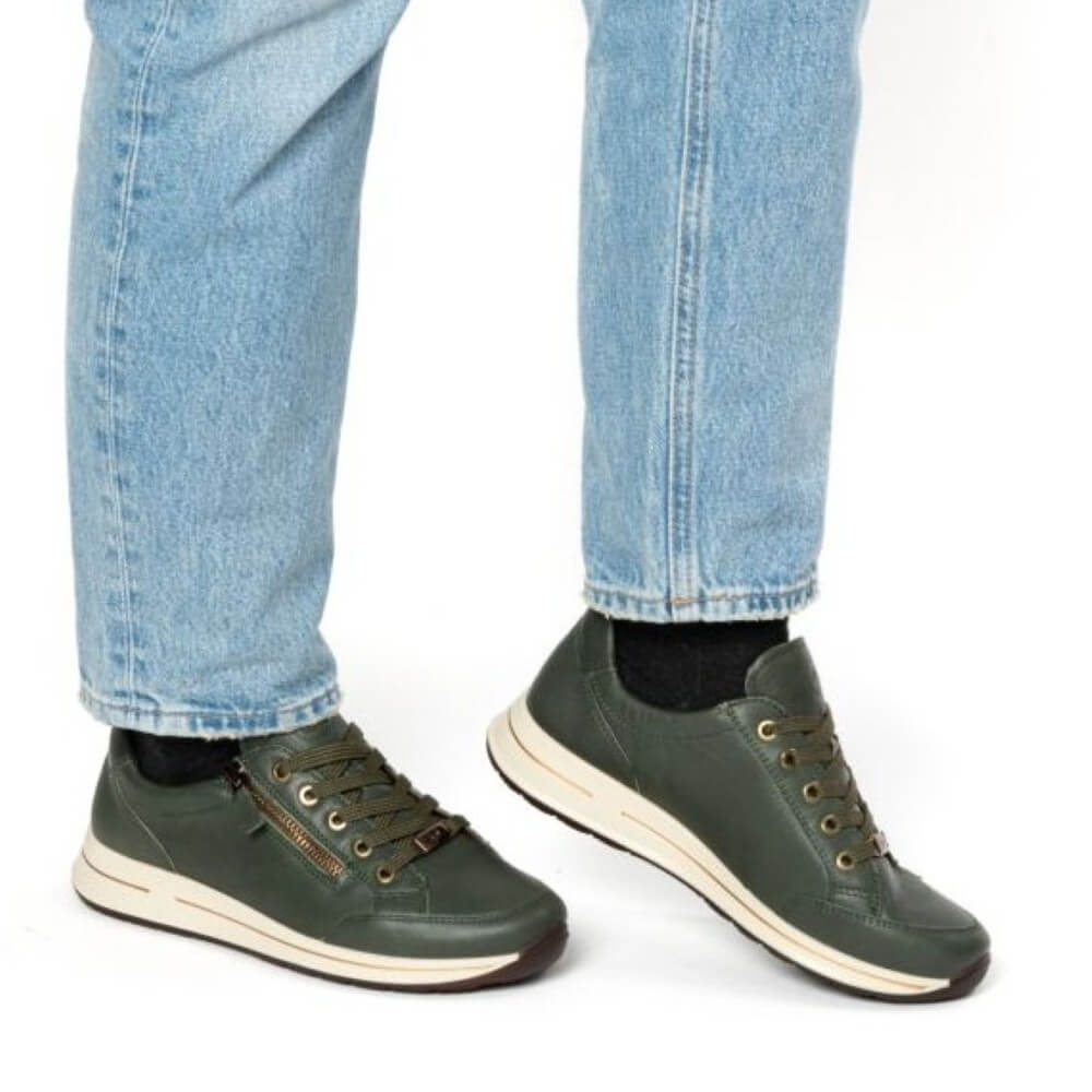A person wearing light blue jeans and dark olive green Ara leather low-top sneakers with cream midsoles, shown from the calves down against a plain white background.