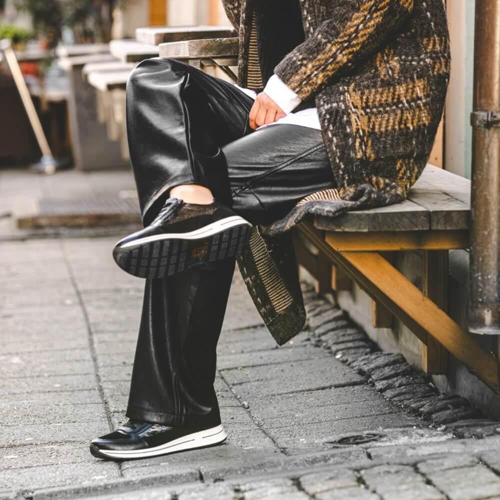 Person wearing black leather shoes and pants sitting on a bench  Black and silver grey sneaker on a white background Ara Osaka 2 Leather Trainers H-Fit 12-24826 Blackoutdoors.