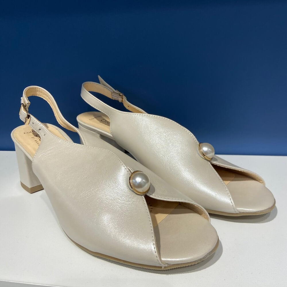 Pair of Bioeco beige open-toe high-heeled shoes with pearl embellishments on a white surface with a blue background.