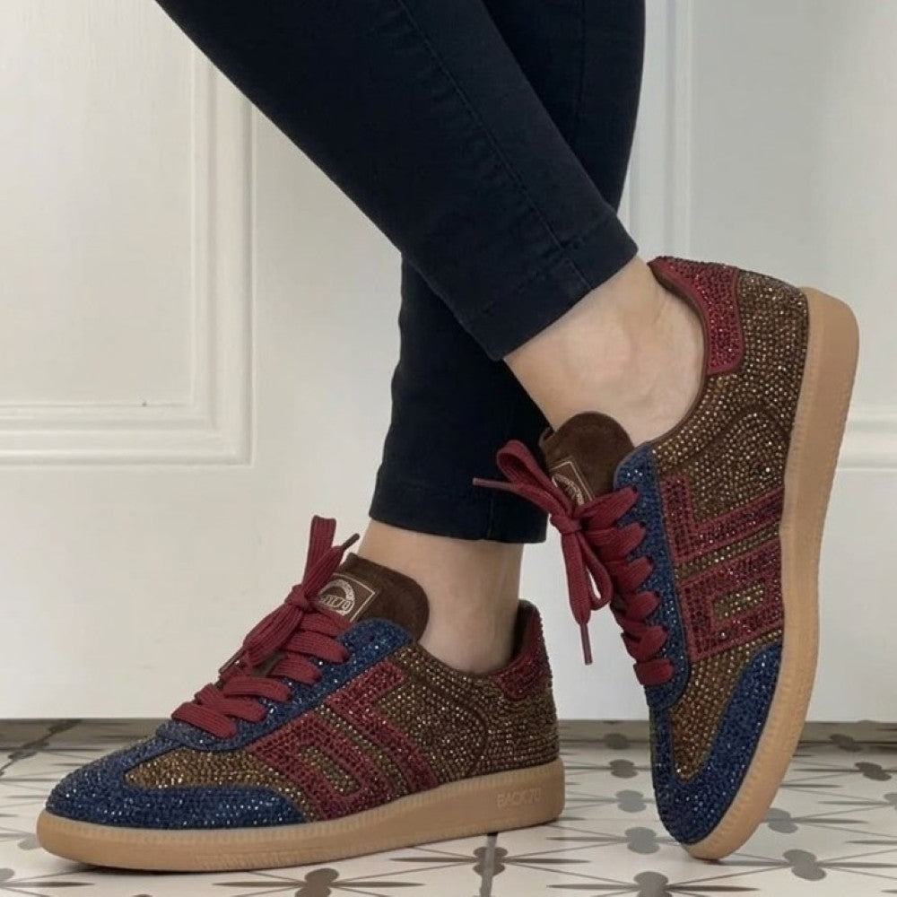 A pair of Back 70 low-top sneakers being worn, shown from the side and back. The shoes feature a brown suede base heavily embellished with gold rhinestones, with dark blue rhinestone panels on the sides and red rhinestone accents on the toe and laces.