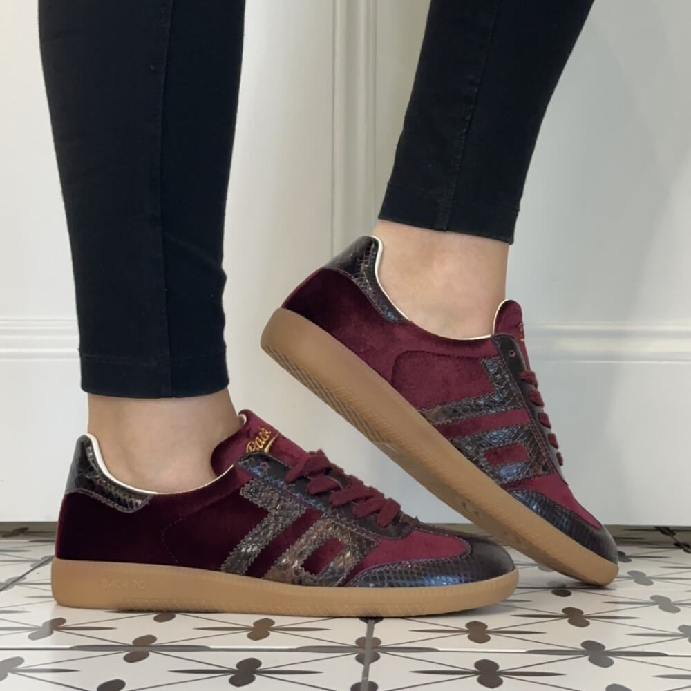 A person wearing Back 70 dark burgundy velvet and snakeskin-patterned  sneakers with tan gum soles, paired with black pants, standing on a patterned tile floor.