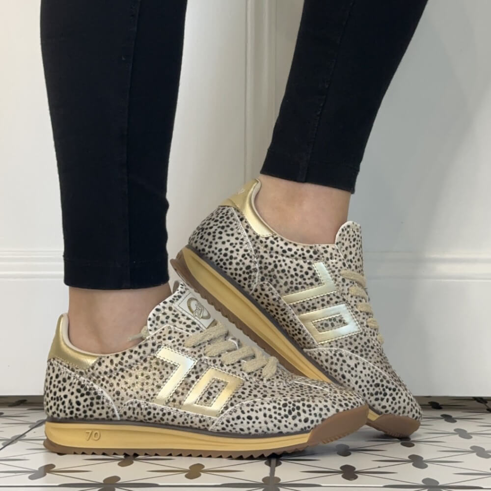 A pair of Back 70 beige and black leopard-print sneakers with metallic gold accents, worn with black leggings on a patterned tile floor.