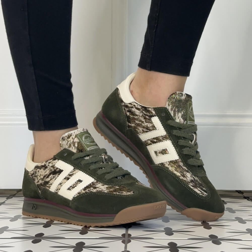 A person wearing dark olive green and beige camouflage Back 70 sneakers with white side stripes, paired with black leggings, standing on a patterned tile floor.