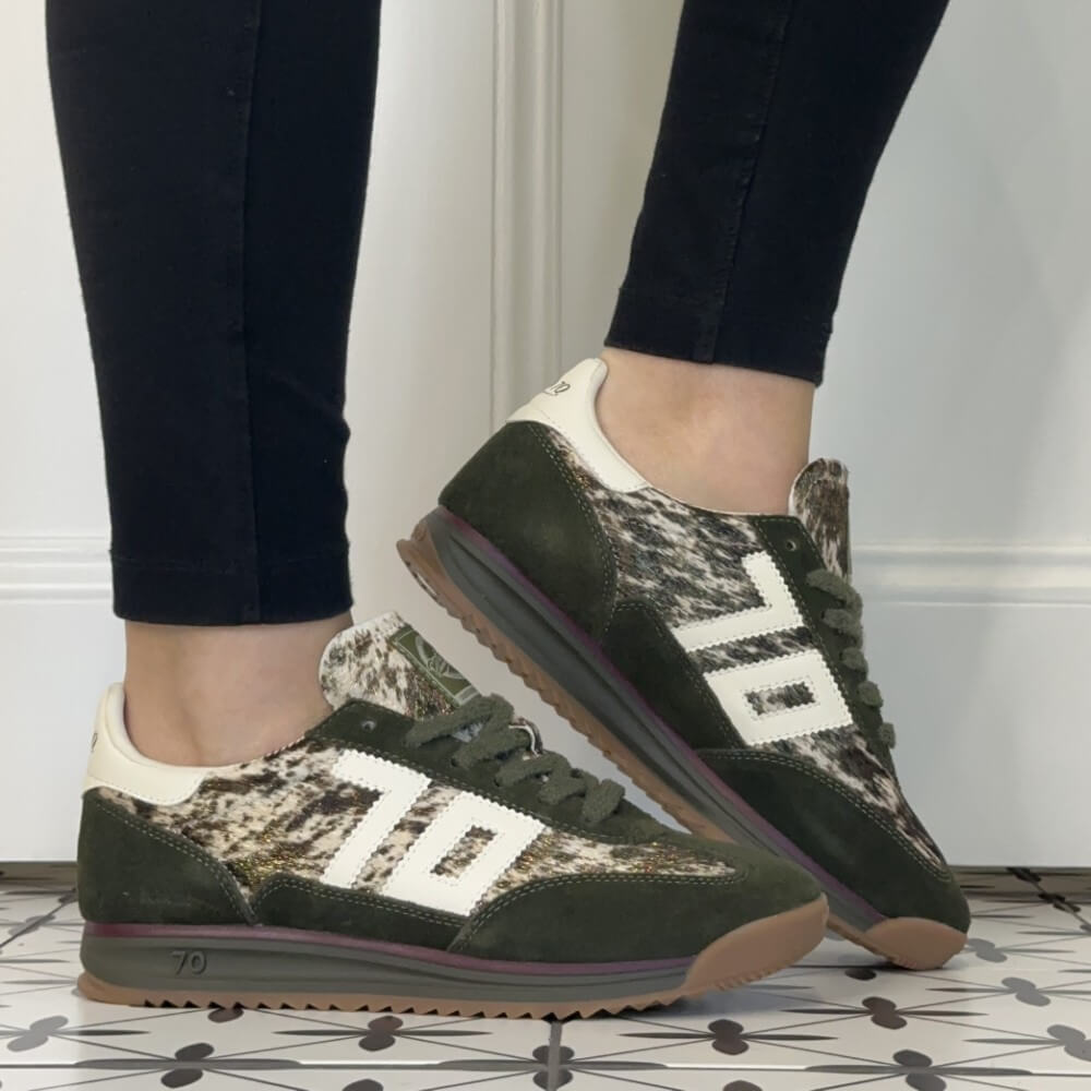 A person wearing dark olive green and beige camouflage Back 70 sneakers with white side stripes, paired with black leggings, standing on a patterned tile floor.