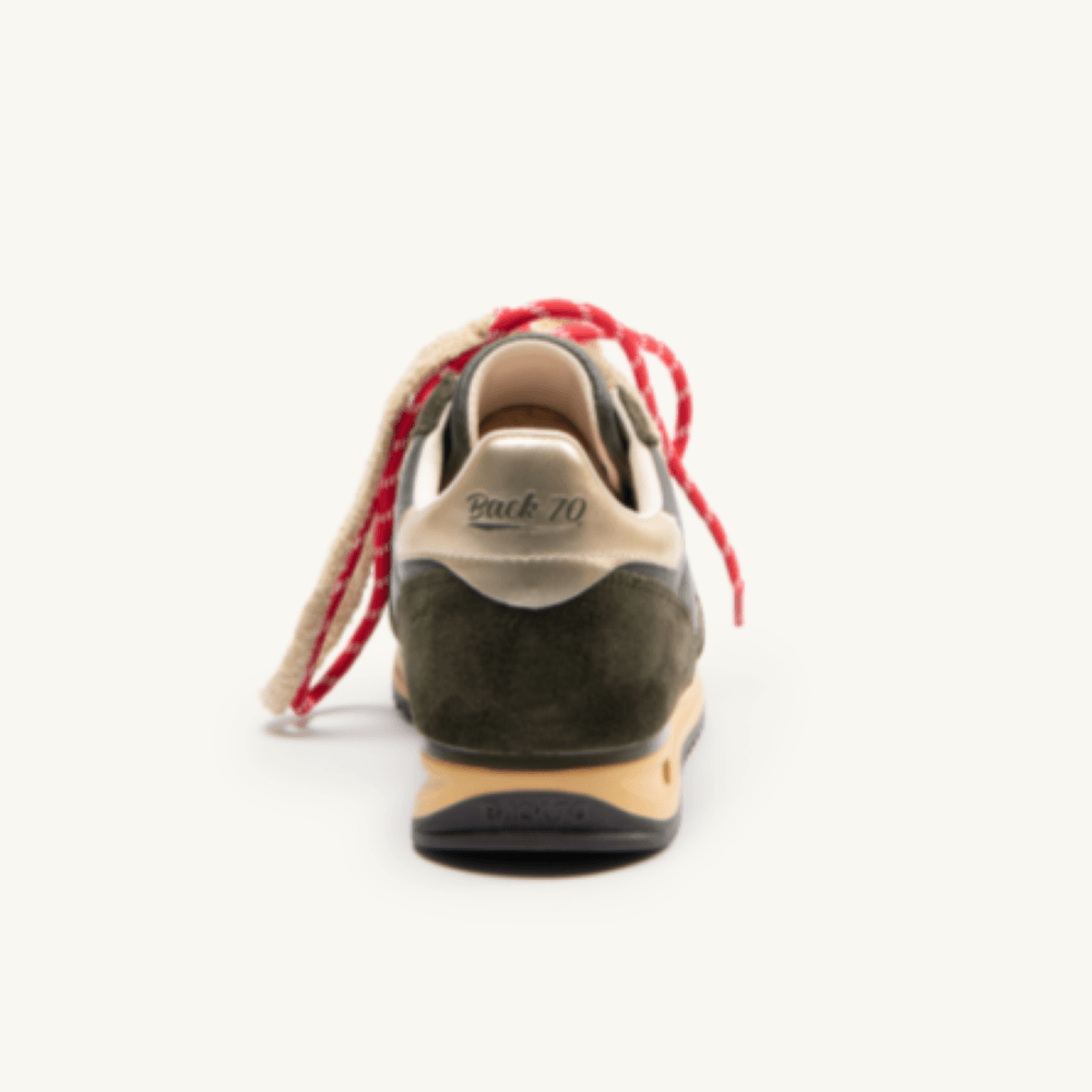 A rear view of a vintage-style sneaker with dark olive green suede, a beige leather heel counter, and distinctive red and cream braided laces, set against a plain off-white background.