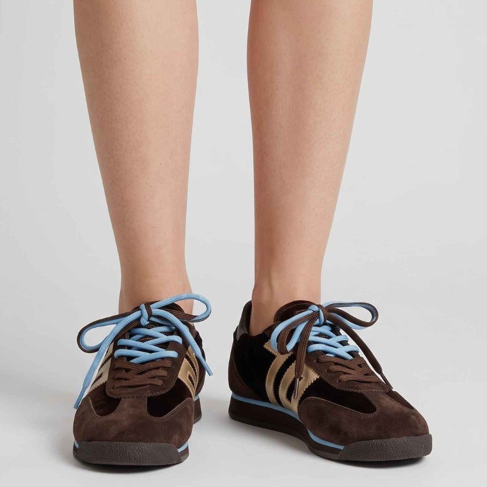 A pair of Back 70 low-top sneakers worn on a model's feet, featuring dark brown suede and mesh panels, light blue laces, and beige side stripes, set against a plain light grey background.