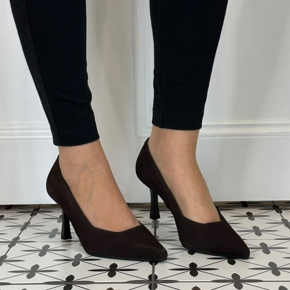 A pair of Bioeco dark brown pointed-toe high heels worn with black skinny jeans, shown on a patterned black and white tile floor against a white wall.