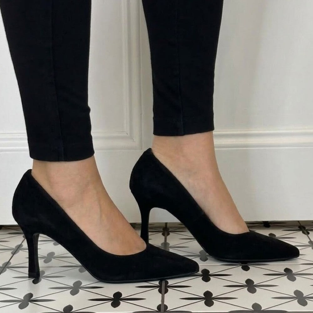 Bioeco Black pointed-toe high heels with slim stiletto heels, shown on a model wearing black pants against a white panelled wall and patterned tile floor.