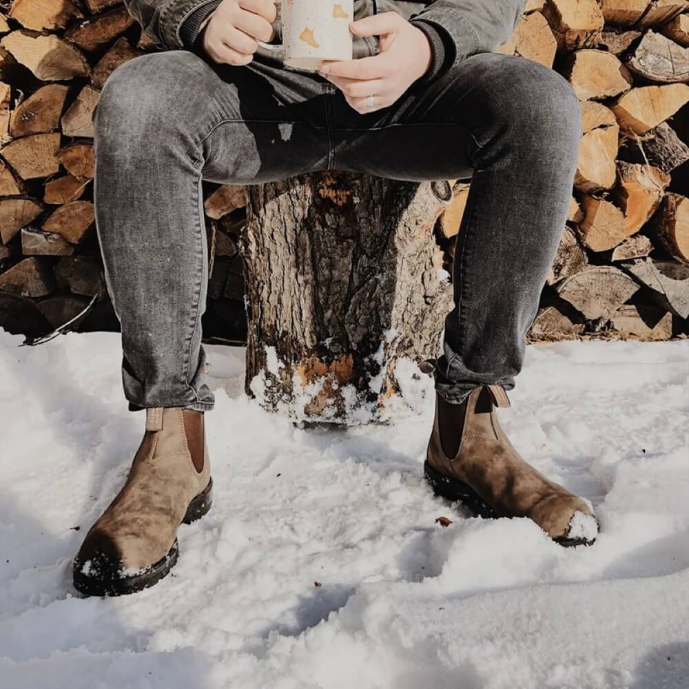 Person sitting on a log in the snow holding a mug, wearing jeans and boots.
