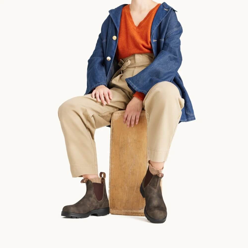 Person wearing a denim jacket, orange shirt, beige pants, and brown boots sitting on a wooden block.