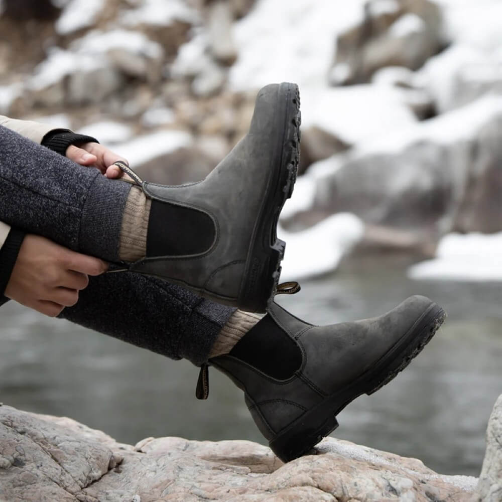 Person wearing black leather Chelsea Boots, sitting on rocks by a body of water.