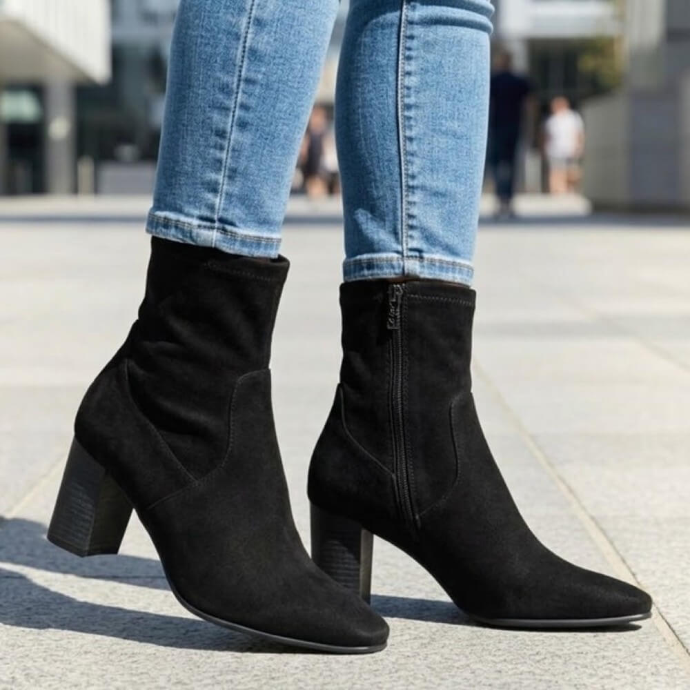 Caprice Black suede ankle boots with block heels and side zippers, shown on a model wearing blue jeans in an outdoor urban setting.