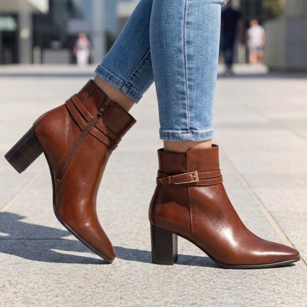 person wearing A Caprice cognac leather ankle boot with a block heel, pointed toe, and a decorative strap with a gold-tone buckle detail around the ankle.
