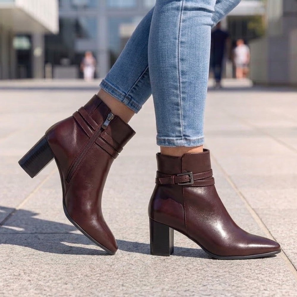 A person wearing Caprice wine leather ankle boot with a block heel, pointed toe, and a decorative strap with a gold-tone buckle detail around the ankle.