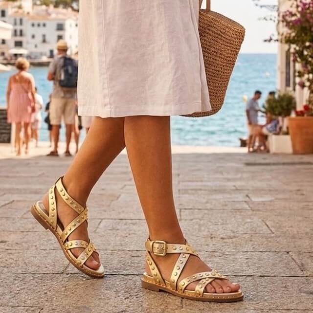 Person wearing Carmela gold sandals with a beach and ocean view in the background