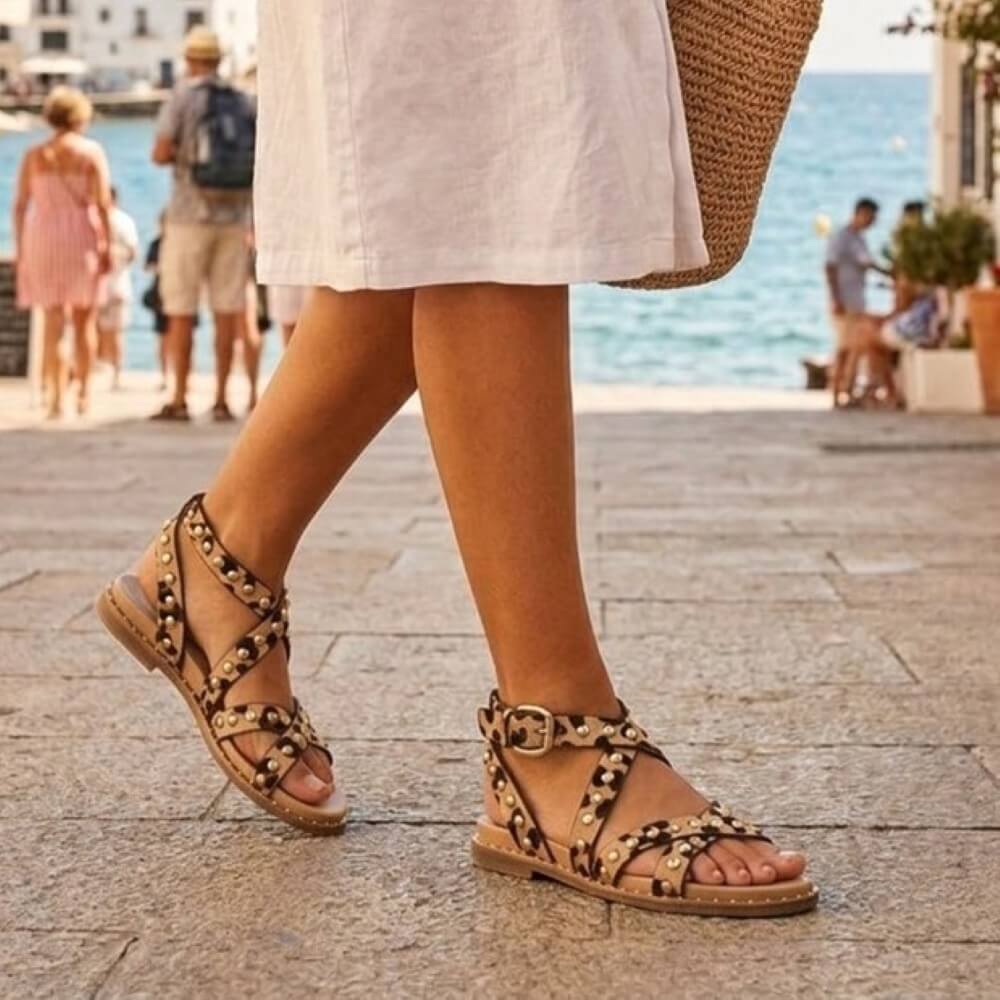 Person wearing brown sandals with decorative straps on a street near the sea.