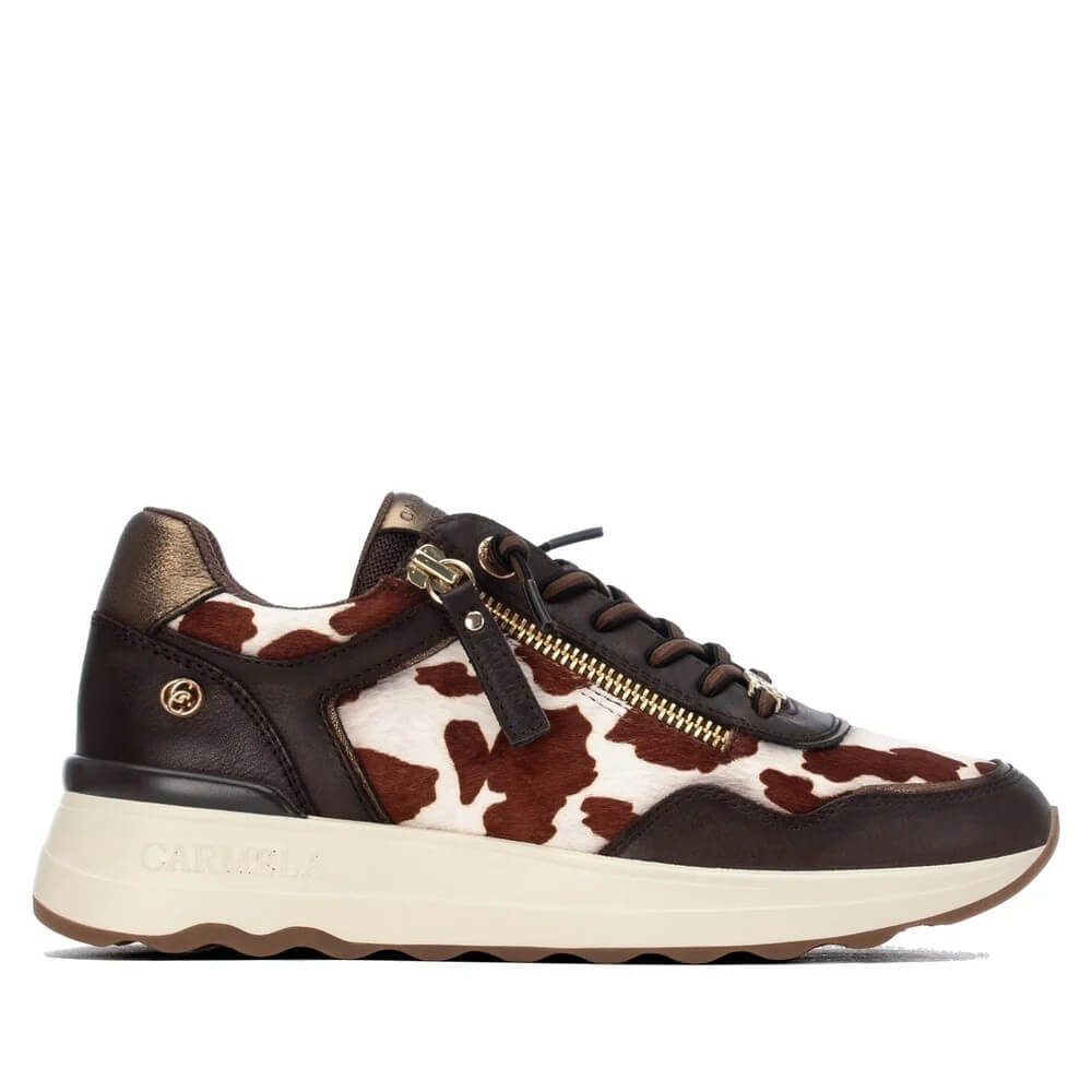 A low-top sneaker by Carmela, featuring a brown and white cow-print pattern, dark brown leather overlays, a gold-tone side zipper, and a thick cream-colored midsole.