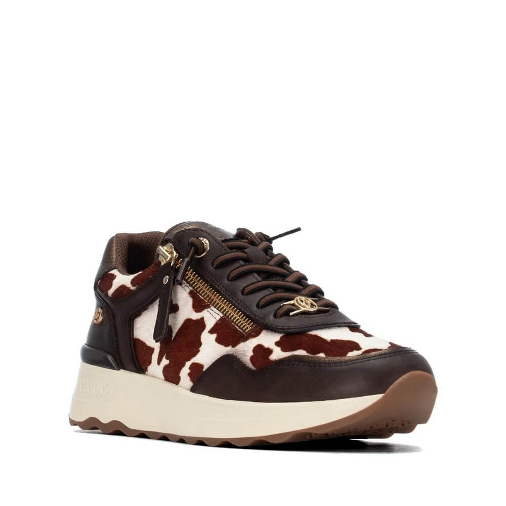 A Carmela low-top sneaker featuring a brown and white cow-print pattern on the upper, dark brown leather overlays, a side zipper with a gold pull tab, and a chunky cream-colored midsole.