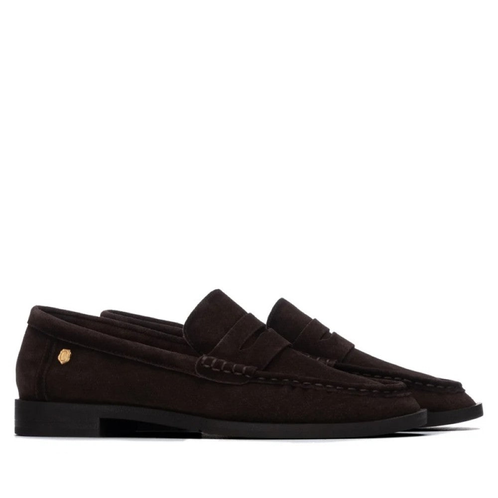 A pair of Carmela dark brown suede penny loafers with black soles and a small gold logo emblem on the heel, shown against a plain white background