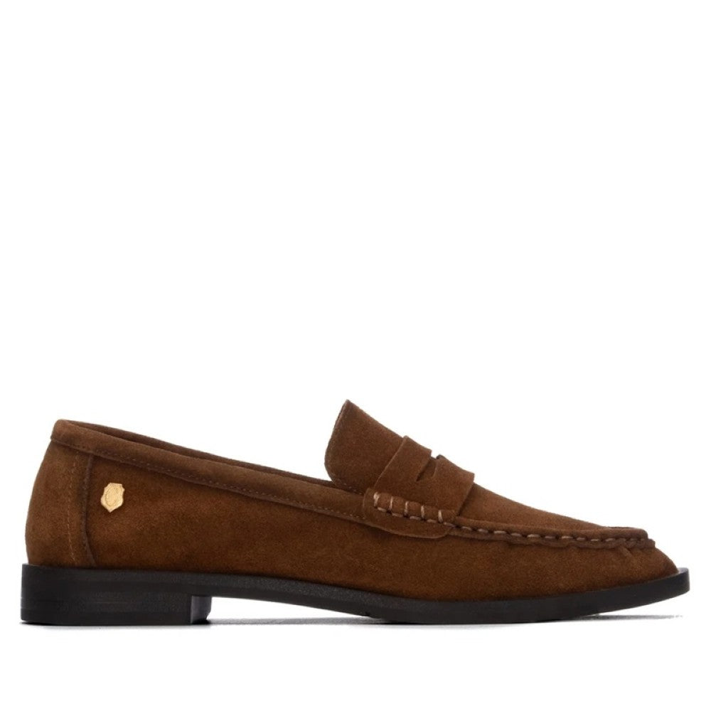 A Carmela brown suede penny loafer shown in profile against a plain white background. The shoe features a small gold-tone shield logo on the heel and a black sole.