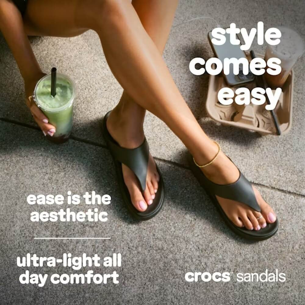 Person wearing black Crocs sandals with a drink, sitting on a concrete surface.
