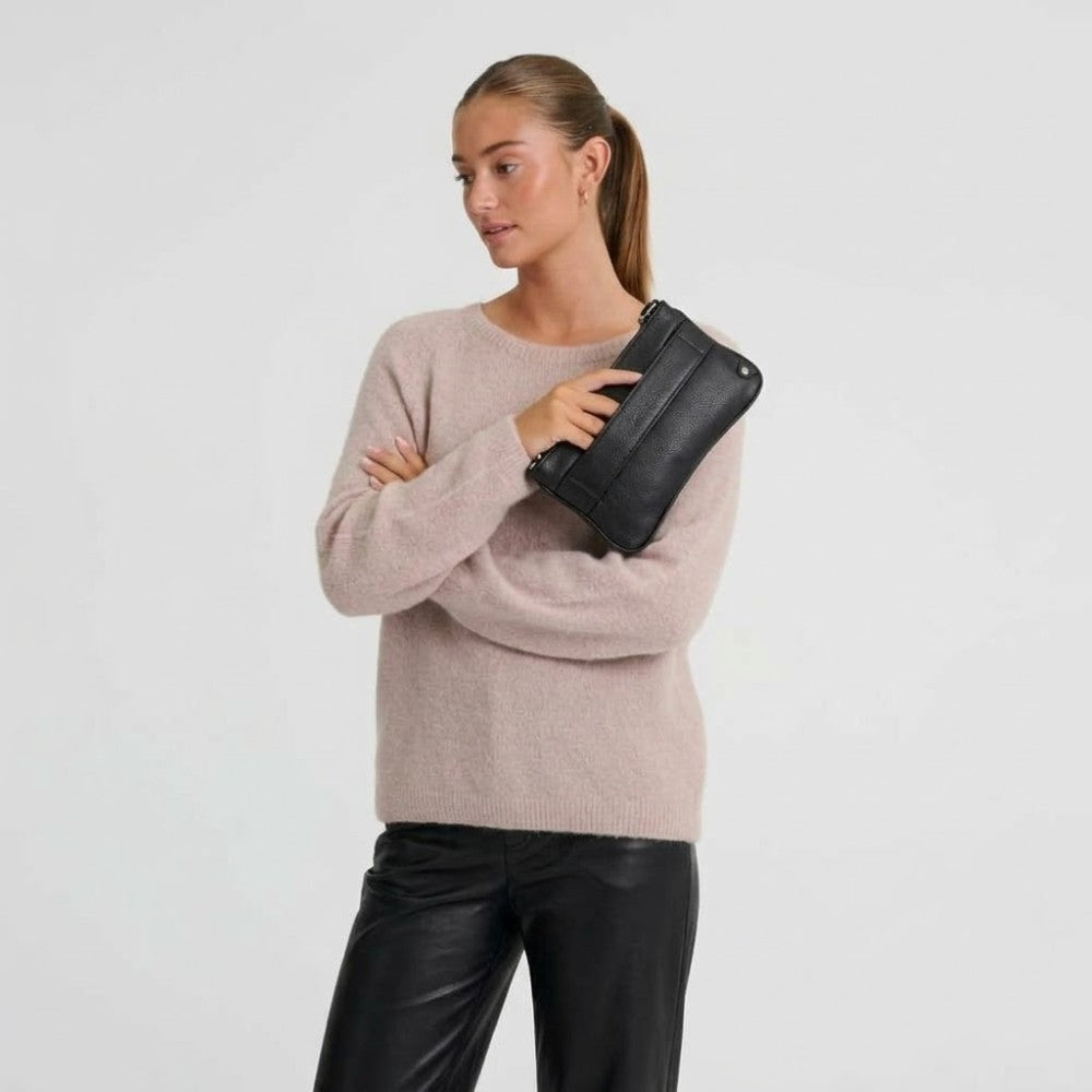 A woman modeling a light pink knit sweater and black leather pants, holding a Depeche black leather clutch bag against her arm, set against a plain light grey background.
