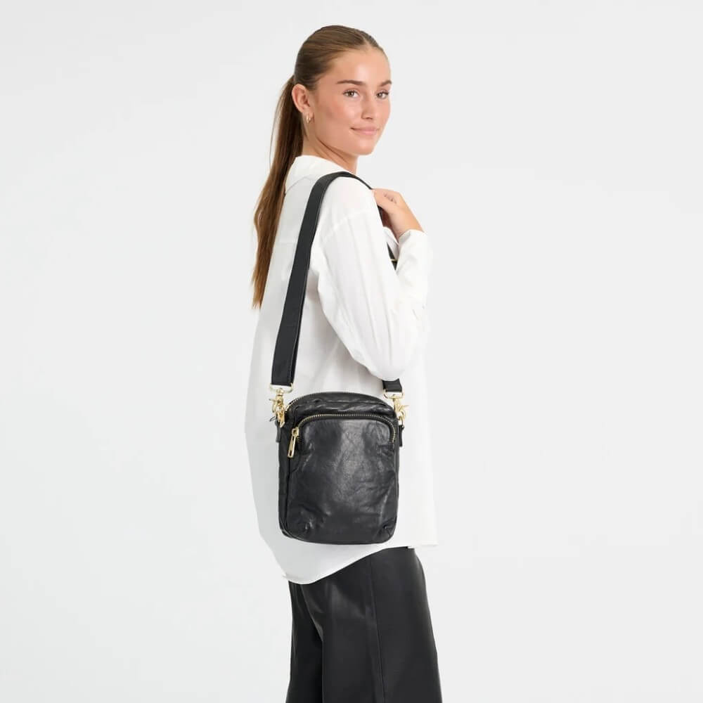 A woman wearing a white long-sleeve shirt and black pants is shown from the side/back, wearing a Depeche black leather crossbody bag with gold-tone hardware and a black strap against a plain white studio background.