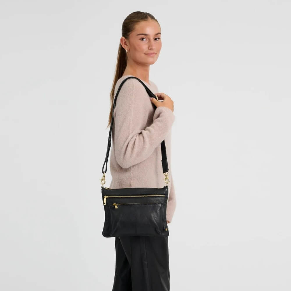 A woman wearing a light pink knit sweater and black pants is shown from the side, wearing a Depeche black leather crossbody bag with gold zippers and hardware against a plain white studio background.