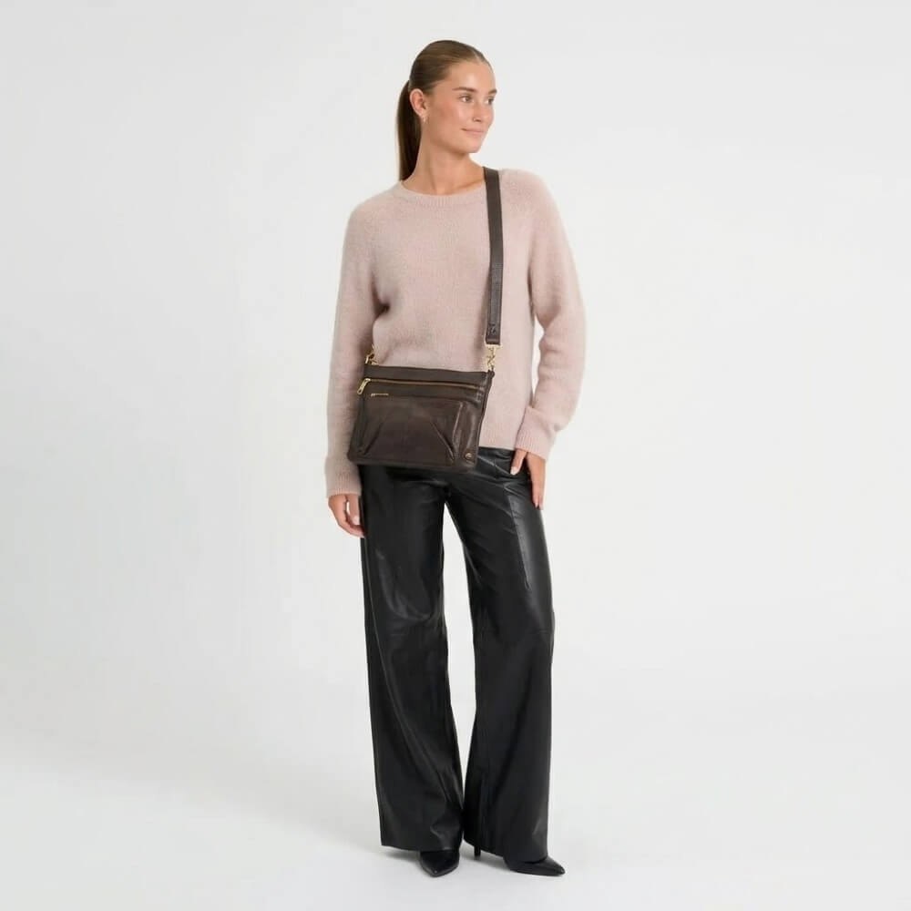 A woman stands against a plain light gray studio background wearing a light blush pink knit sweater, black wide-leg leather pants, and black pointed-toe heels, while carrying a dark brown crossbody bag with gold-tone hardware.