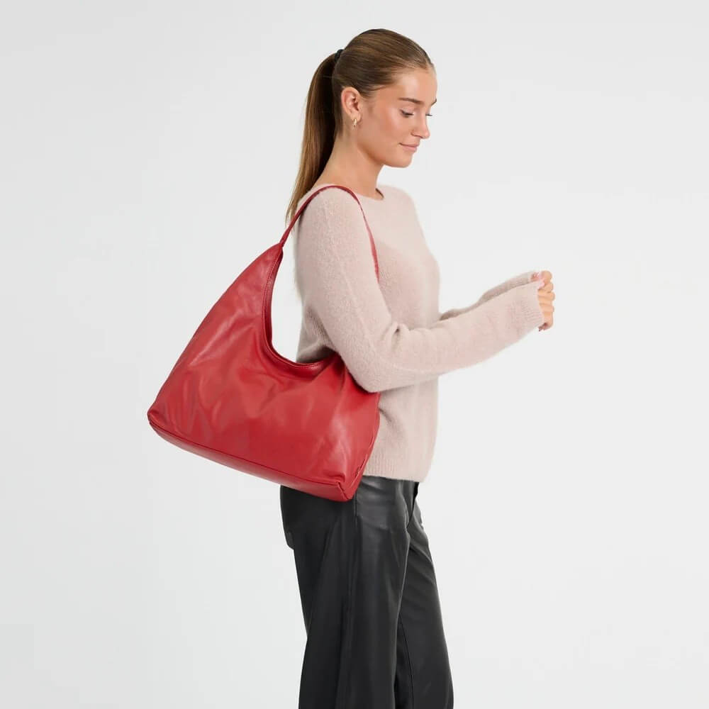 A woman in a light pink sweater and black pants carries a large Depeche red leather shoulder bag against a plain white studio background.