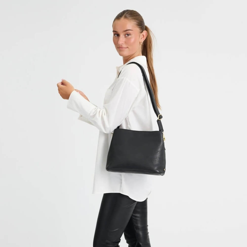 A woman wearing a white button-up shirt and black leather pants carries a Depeche black leather shoulder bag with a gold-tone zipper pull against a plain white studio background.