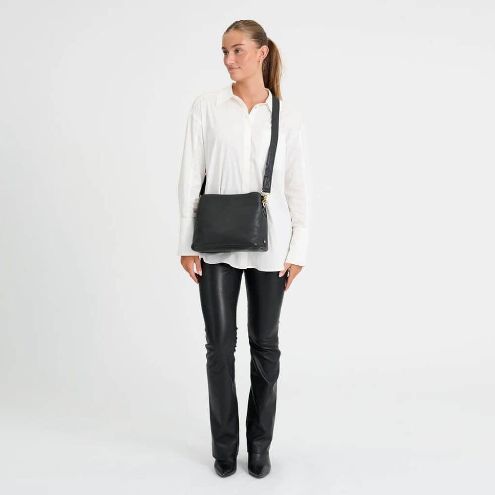 A woman modelling a Depeche black leather crossbody bag with a gold zipper, worn across her torso over a white button-down shirt and black pants, set against a plain white studio background.