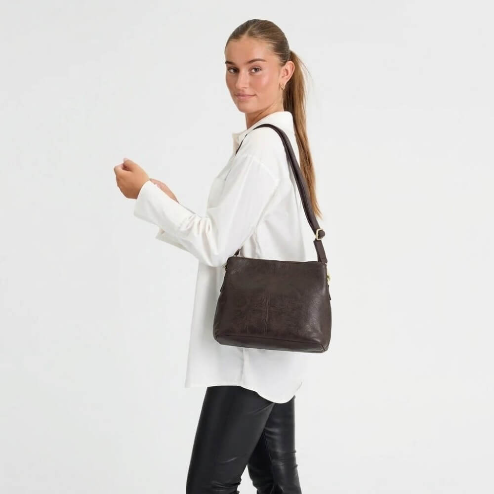 A woman wearing a white button-up shirt and black leather pants carries a Depeche Brown leather shoulder bag with a gold-tone zipper pull against a plain white studio background.