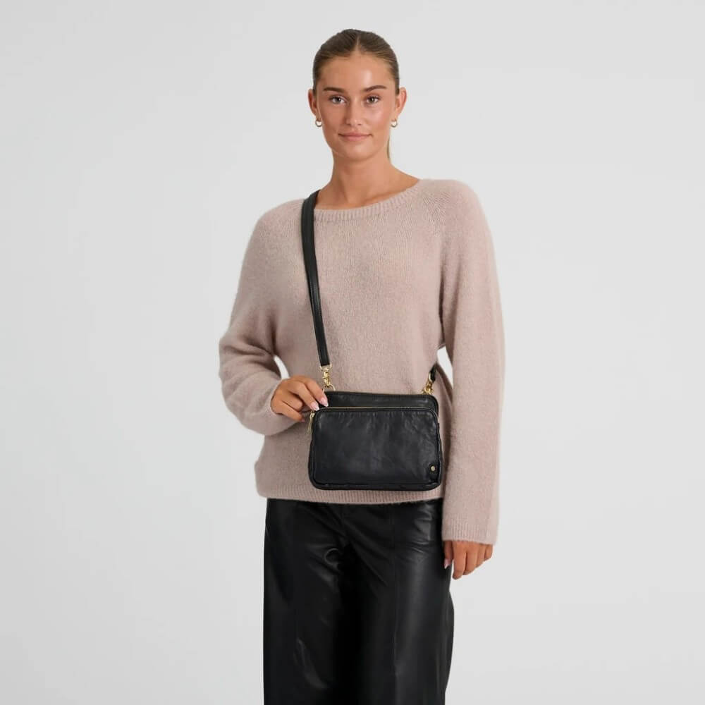 A woman wearing a light pink knit sweater and black leather pants models a Depeche black leather crossbody bag with gold-tone hardware against a plain light grey background.