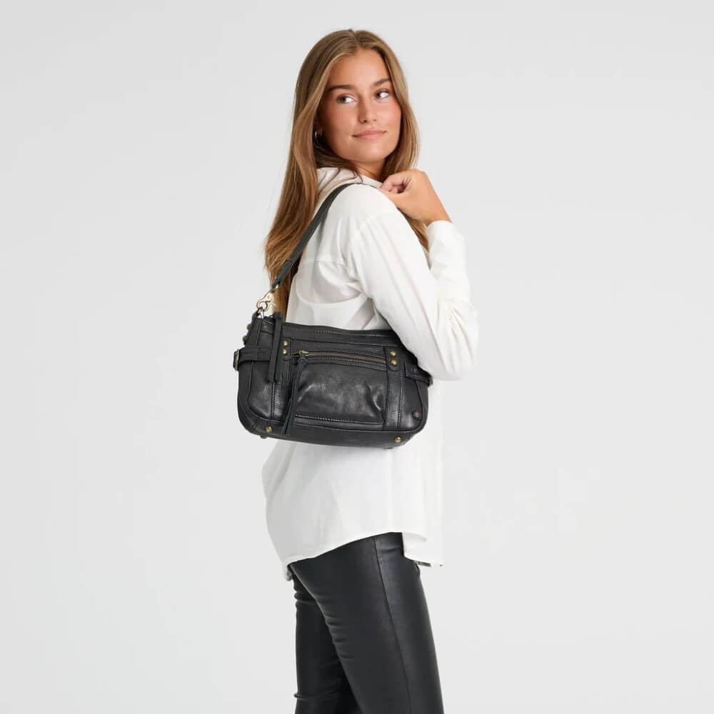 A woman wearing a white long-sleeve shirt and black pants carries a Depeche black leather crossbody bag with gold-tone hardware against a light grey studio background.