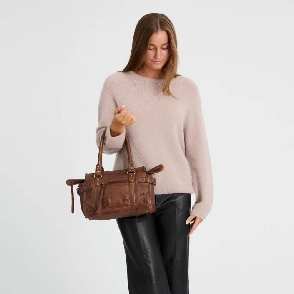A woman wearing a light pink knit sweater and black leather pants holds a Depeche brown leather handbag against a plain light grey studio background.