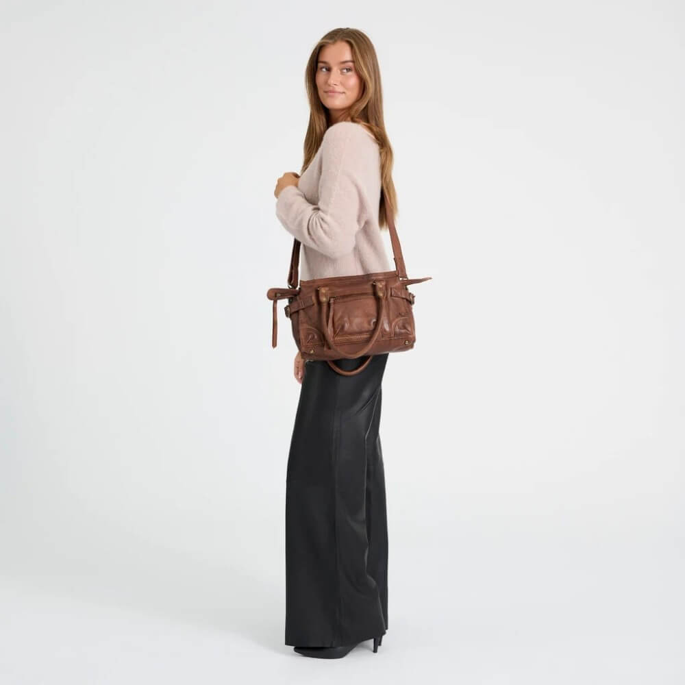 A woman wearing a beige sweater and black wide-leg pants carries a Depeche brown leather shoulder bag with multiple front pockets, photographed against a clean white studio background.