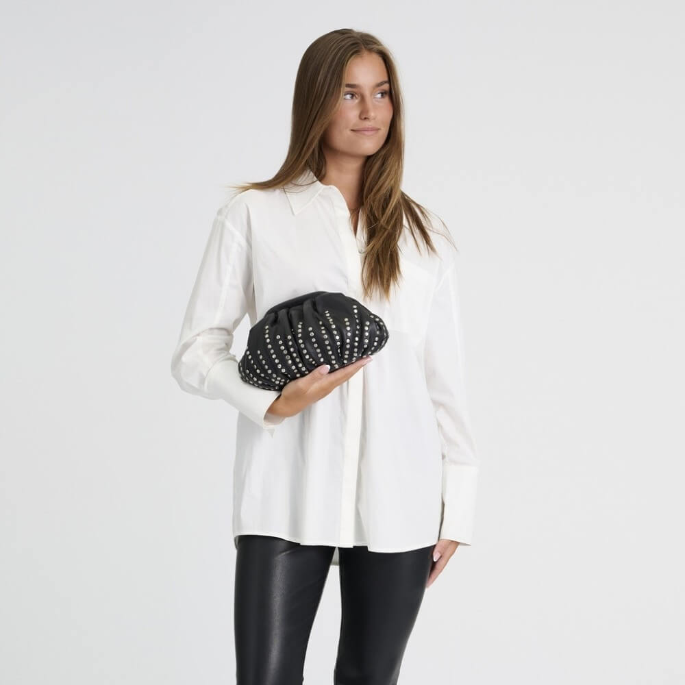 A woman modelling a white long-sleeve button-up shirt and Depeche black leather pants, holding a black clutch bag with silver studs against a plain light grey background.