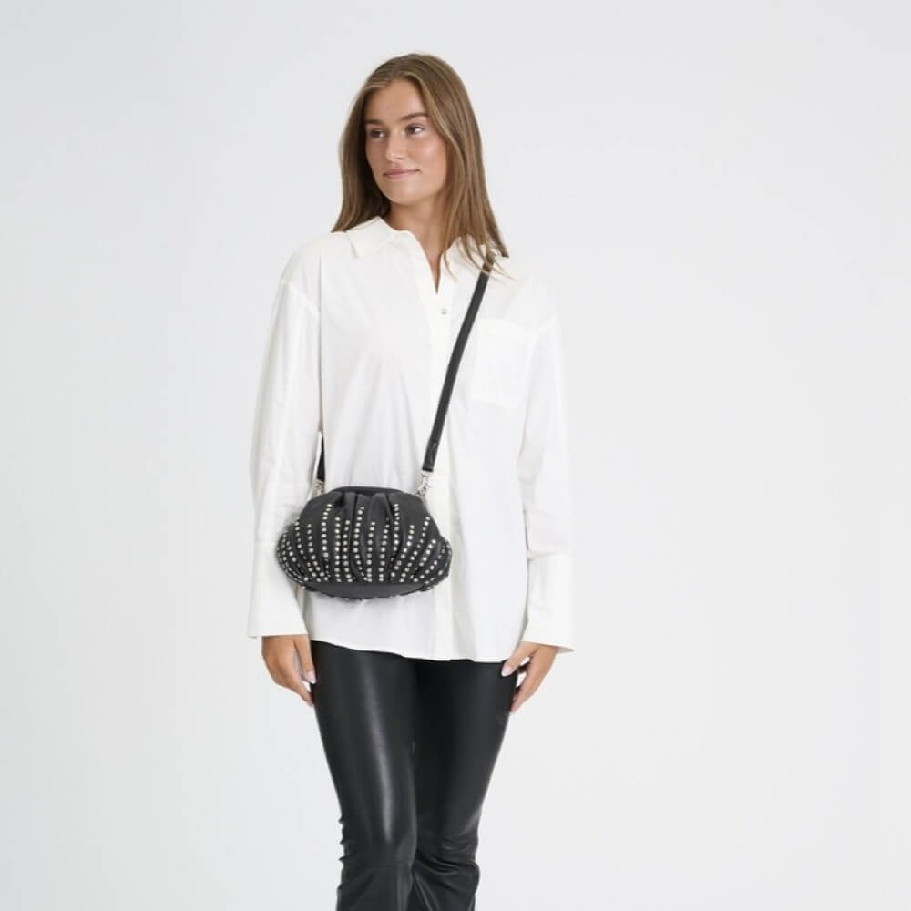 A woman wearing a white button-down shirt and black pants models a Depeche black studded crossbody bag with a thin black strap against a plain white studio background.