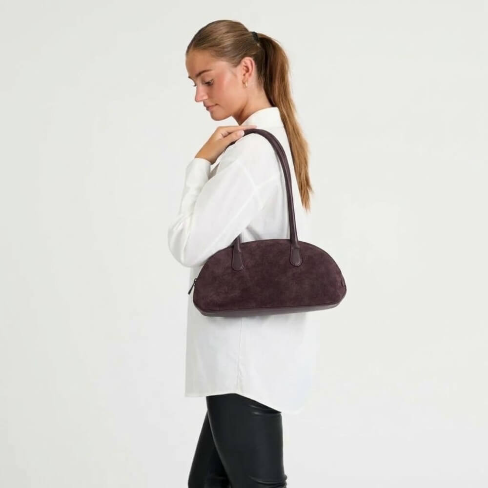 A woman in a white button-up shirt and black pants carries a Depeche Plum suede dome-shaped shoulder bag against a plain white studio background.