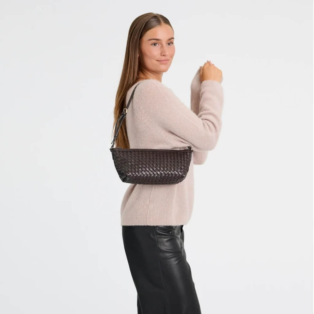 A woman wearing a pink jumper and black pants holds a Depeche dark brown woven-texture shoulder bag with a thin brown strap against a plain light grey studio background.