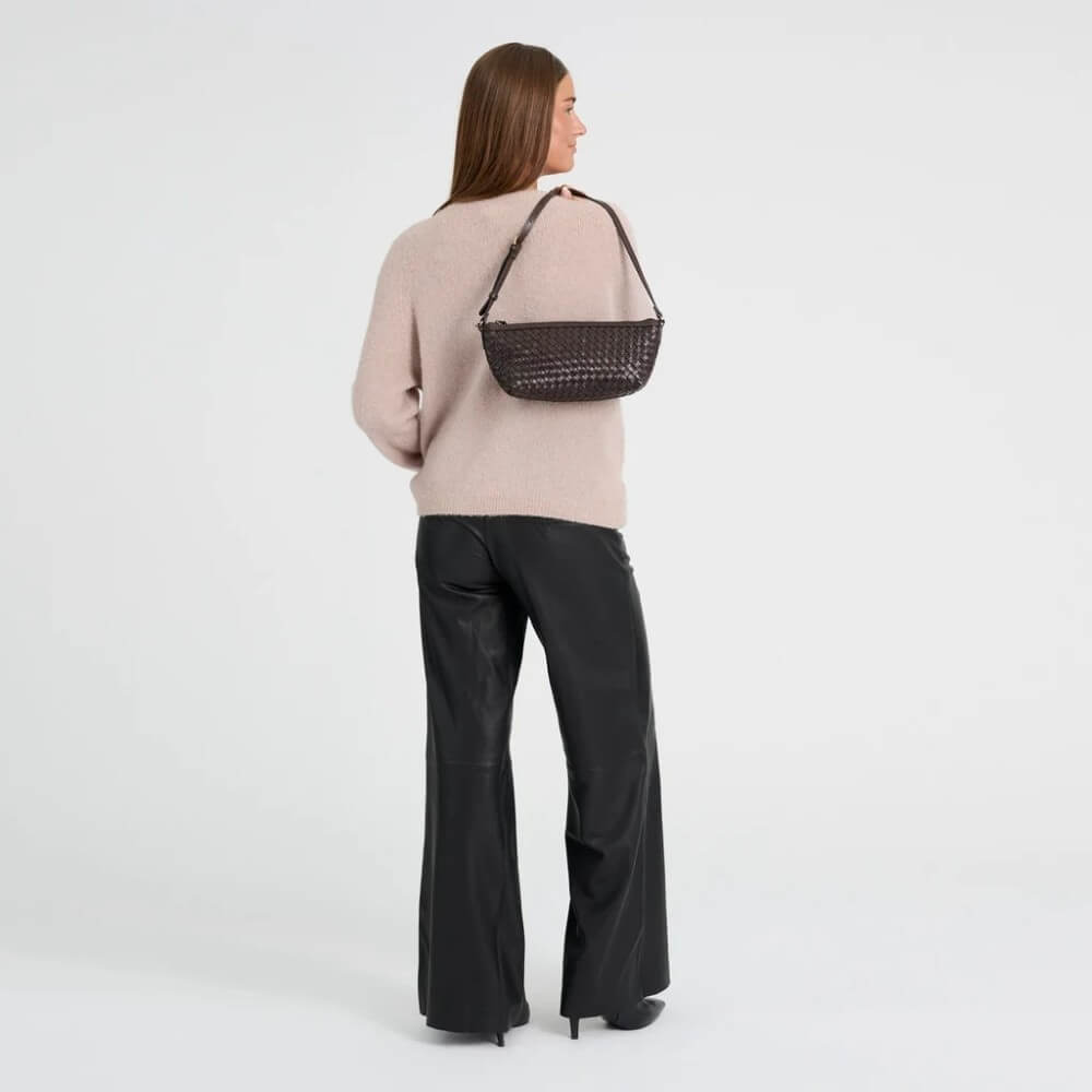 A woman wearing a pink jumper and black pants holds a Depeche dark brown woven-texture shoulder bag with a thin brown strap against a plain light grey studio background.