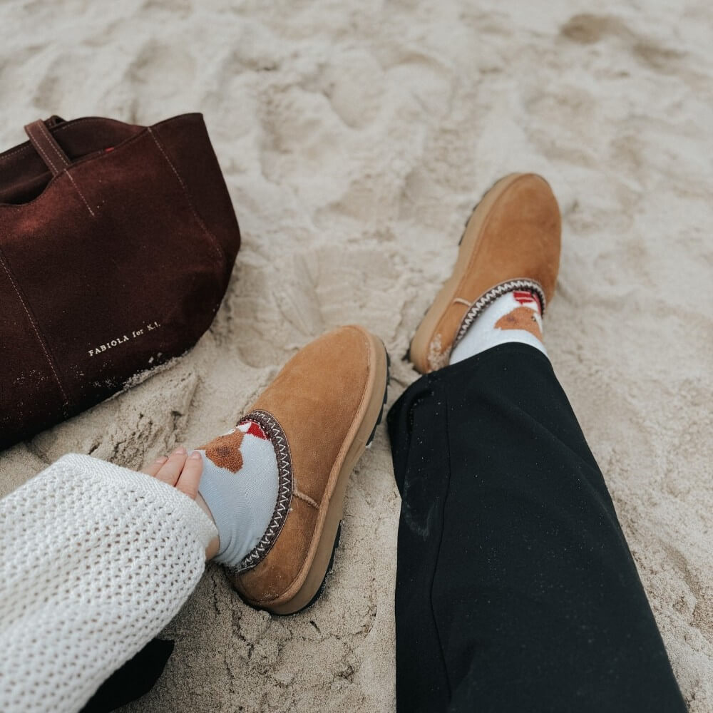 Person wearing brown moccasin-style shoes on a sandy surface with a dark brown bag nearby. EMU Sharky Scuff Chestnut