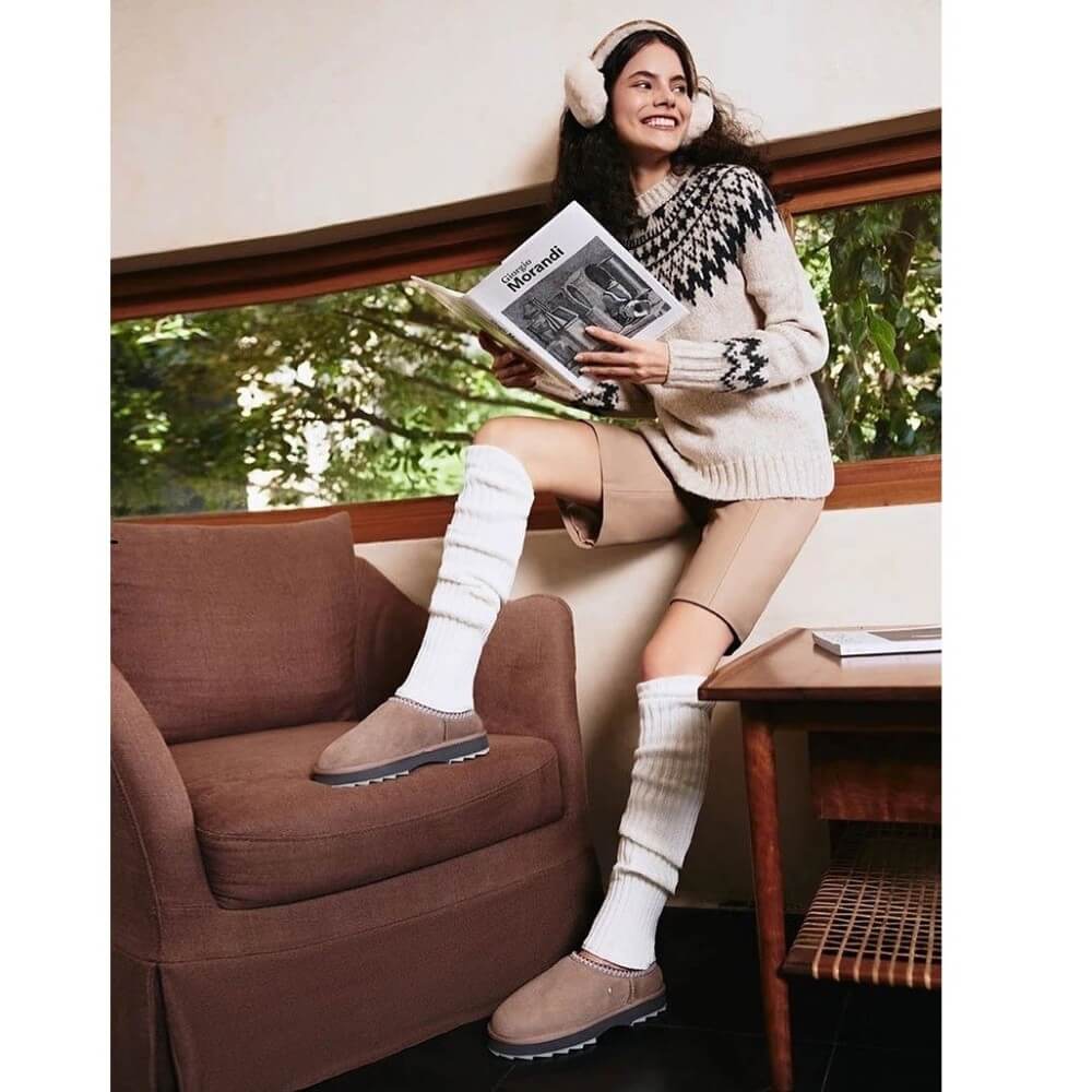 Woman sitting on a couch reading a magazine, wearing a patterned sweater, knee-high socks, and earmuffs. EMU Sharky Scuff Chestnut